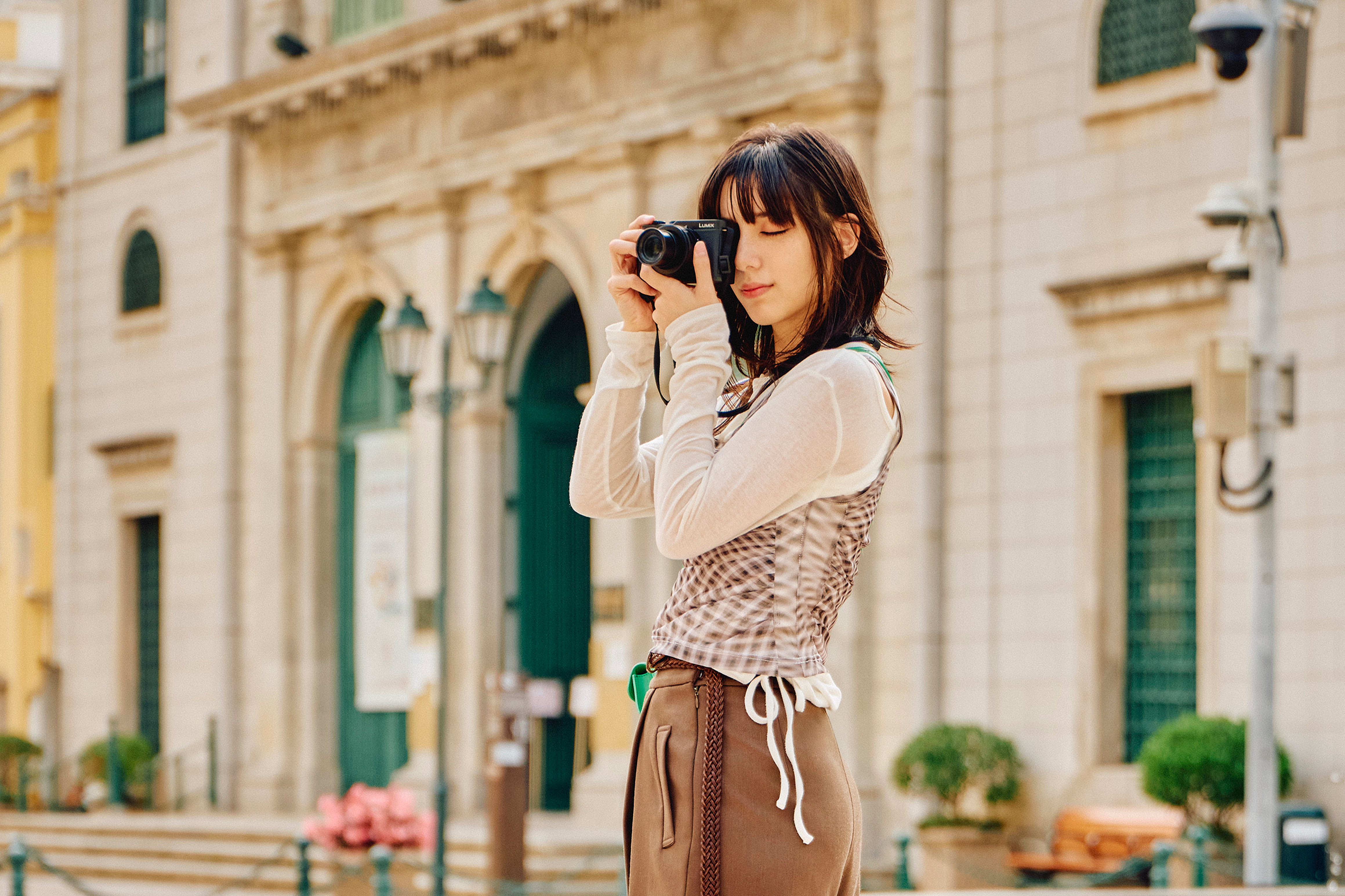 A woman stands in front of a building, looking through the Lumix L10 camera she holds to her eye