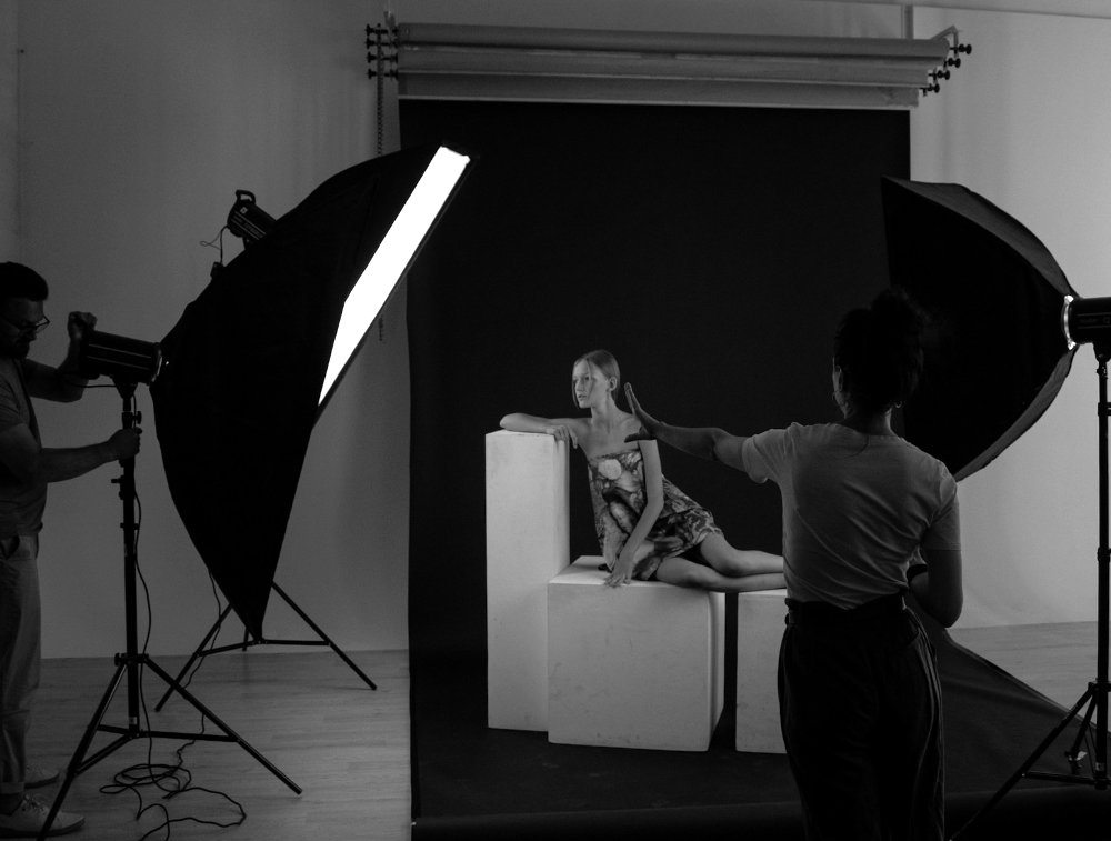 Black and White Photograph: A model on boxes against a backdrop, surrounded by lit softboxes. In the foreground, the photographer directs their assistant to adjust a light.