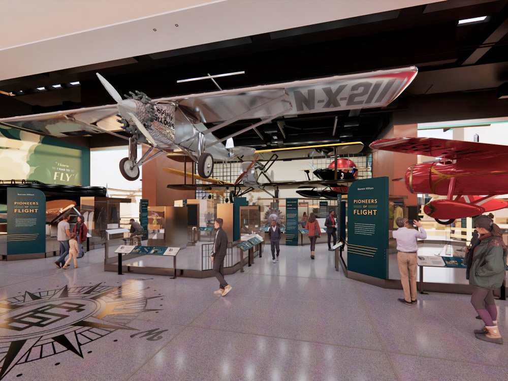 An artist's concept of the Barron Hilton Pioneers of Flight exhibition. In the center is a hanging silver airplane, a red airplane peaks out of the right. There are vertical panels with information on it.
