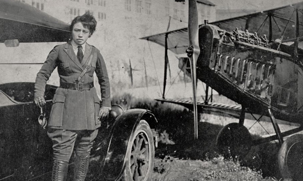 Bessie Coleman standing by a car and airplane, wearing a pilot's outfit, with a building in the background.
