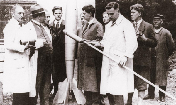 A historical black and white photograph showing a group of individuals examining an early model rocket outdoors.