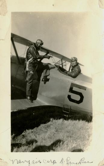 Two pilots in vintage flight gear interact beside a small biplane marked with the number 5 and labeled Naval Air Corps Reserve.