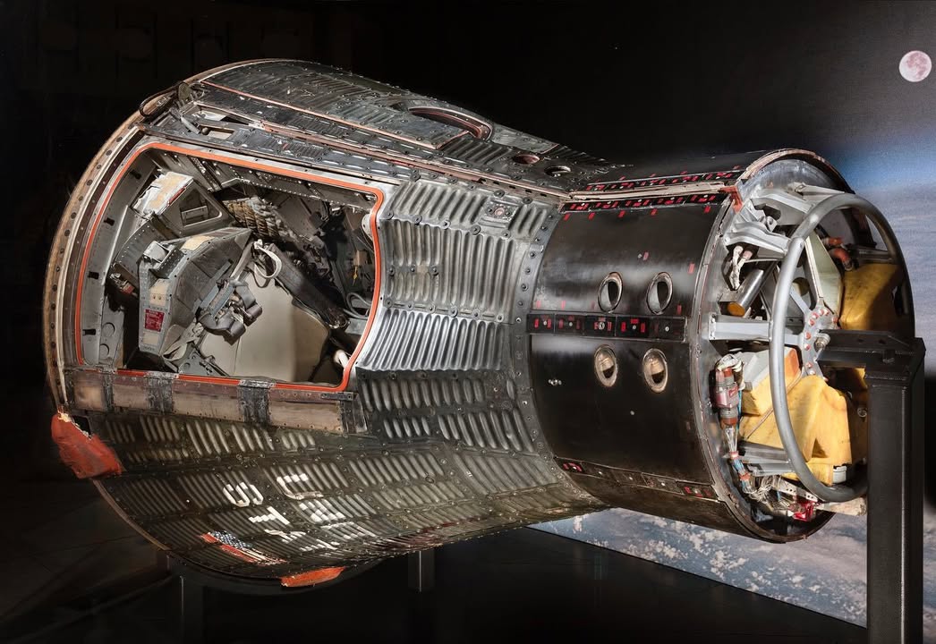 Display of a Gemini spacecraft, shown on its side, with exposed interior including control panels and seats, against a dark background.