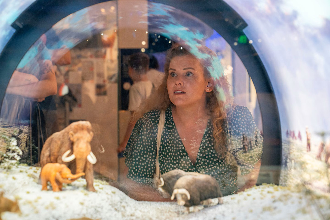 a woman with long, wavy blond hair looks into a diorama in the Fossil Hall