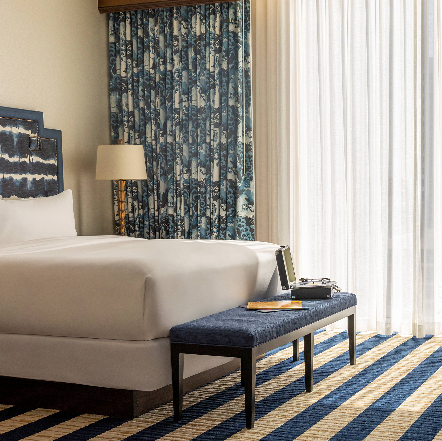 Guest room with a bed, chair, upholstered bench, lamps, rug and curtains decorated in cream and blue colors at the Fairmont Austin hotel in Downtown Austin, Texas