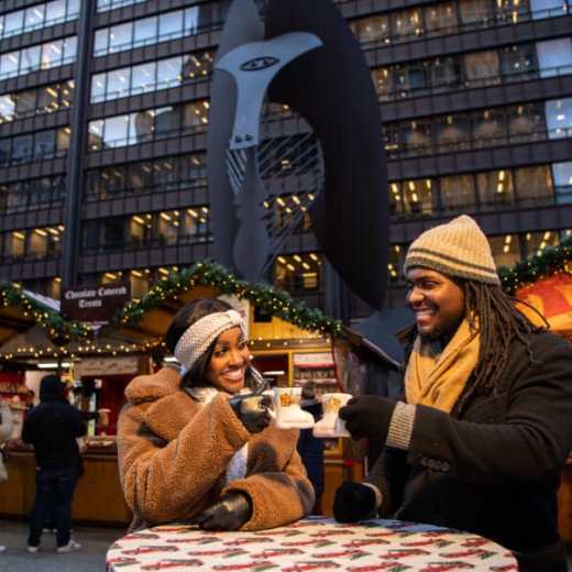 A woman and man toast with boot-shaped mugs against a backdrop of festively decorated wooden booths at Christkindlmarket in Chicago's Daley Plaza