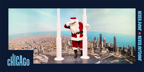 A person wearing a Santa Claus suit stands between two white poles at the top of Willis Tower and peers down at the city of Chicago