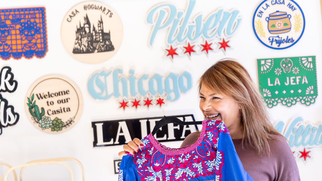 A woman holds up a colorful blue-and-red blouse with a mural promoting the Pilsen neighborhood behind her