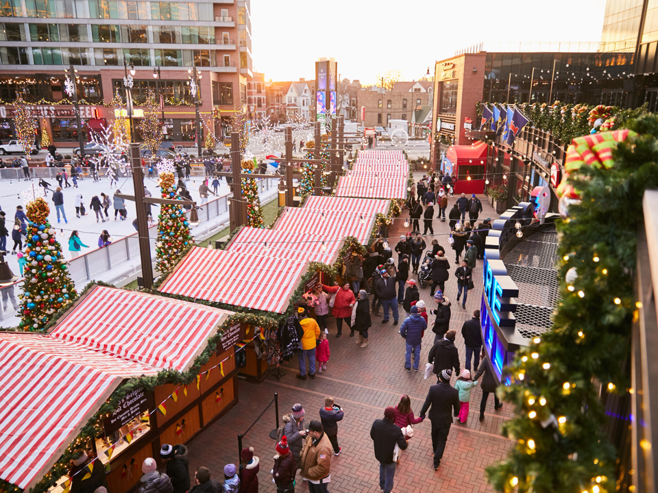 An aerial view of Christkindlmarket Wrigleyville, with red-and-white striped vendor booths and skaters on an ice rink in the background