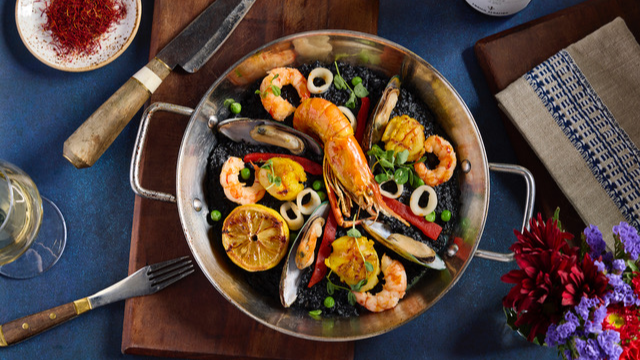 Overhead view of a pan filled with a seafood paella-style dish featuring shrimp, mussels, squid rings, lemon halves, and vegetables on a blue tabletop, with a knife, wine glass, and flowers nearby.