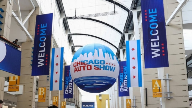 The grand concourse in McCormick Place with a hanging blue globe sign reading ‘Chicago Auto Show,’ surrounded by vertical ‘Welcome’ banners and Chicago flag–themed displays.