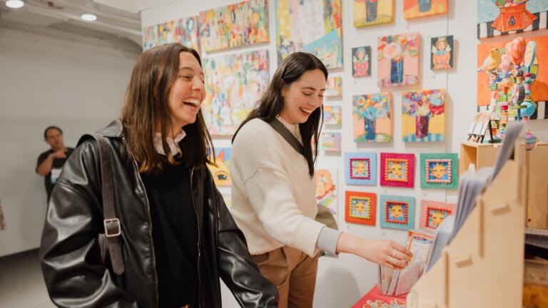 Two women laughing while perusing merchandise at the One of a Kind Show