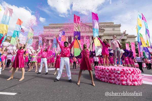 Performers in bright pink outfits dance and toss confetti into the air as colorful banners wave, with the National Archives building serving as a grand backdrop for the Cherry Blossom Festival parade.