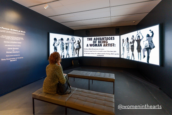 A visitor sits on a bench, gazing at a large, black-and-white digital display titled “The Advantages of Being a Woman Artist,” inside the National Museum of Women in the Arts.