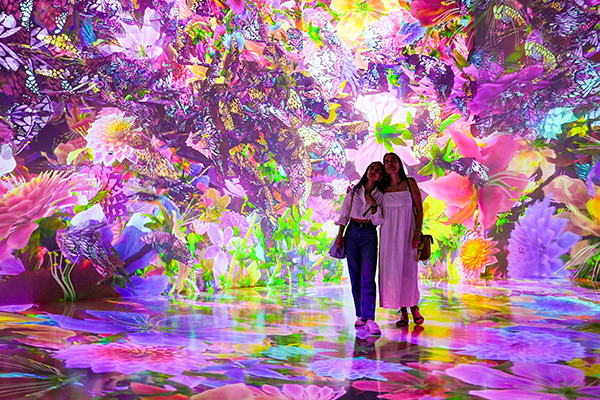 Two women stand inside an immersive digital art installation, surrounded by vibrant, oversized projections of colorful flowers covering the walls and floor.
