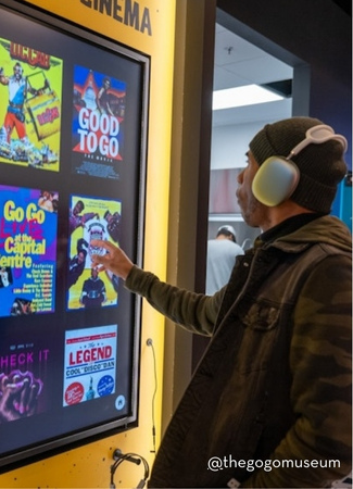 A visitor wearing headphones interacts with a touchscreen display showcasing Go-Go music history, featuring colorful posters and album covers at the all-new Go-Go Museum & Cafe in DC.