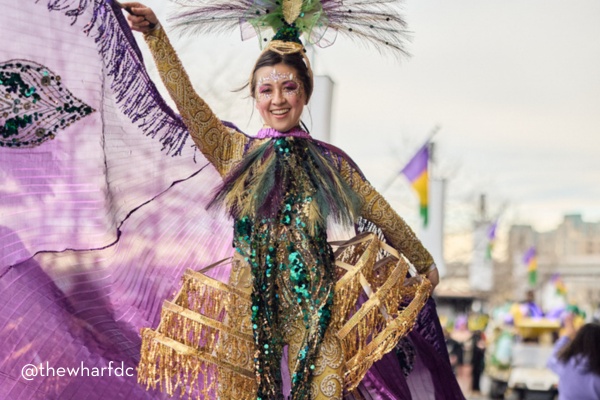 A performer in a vibrant gold and green sequined outfit, with a feathered headdress and purple cape, dances joyfully during a Mardi Gras celebration at The Wharf, with festive flags waving in the background.