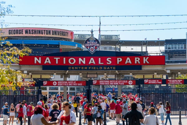 Fans dressed in red gather outside the entrance of Nationals Park under a bright blue sky, with the stadium’s signage and championship banners proudly displayed above.