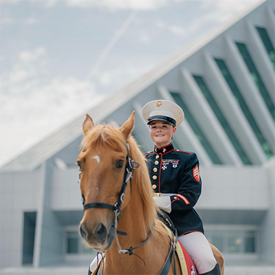 A U.S. Marine in full dress uniform sits atop a chestnut-colored horse, smiling confidently in front of a modern angular building with glass windows.