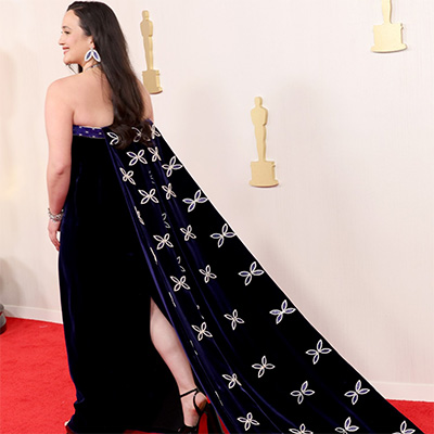 A woman with long dark hair walks the red carpet wearing a deep blue velvet gown with an embroidered cape featuring white floral designs, standing against a backdrop with gold Oscar statuettes.