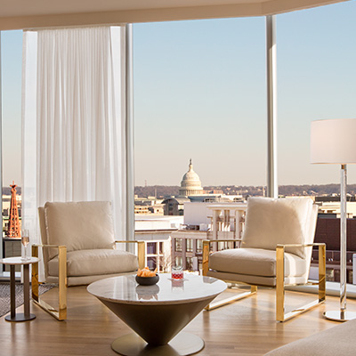 A modern hotel lounge with sleek white chairs, a round coffee table and floor-to-ceiling windows offering a view of the U.S. Capitol in the distance.