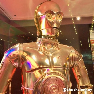  A close-up of the golden C-3PO figure from Star Wars, reflecting colorful lights in a museum setting.