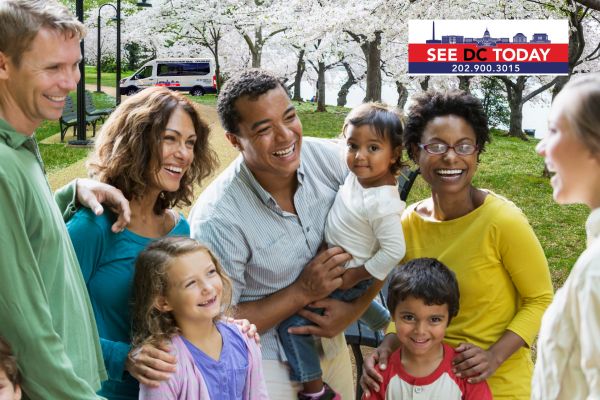 A diverse group of smiling people, including children, gather under blooming cherry blossoms in a park, with a tour van in the background and a 'See DC Today' logo in the top right corner.