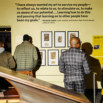 Visitors in a museum gallery observe framed artwork on a yellow wall beneath a quote by artist Elizabeth Catlett.