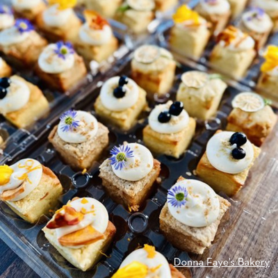 An assortment of beautifully decorated miniature pastries topped with fresh fruit, edible flowers, and cream, arranged in clear plastic trays on a wooden surface.