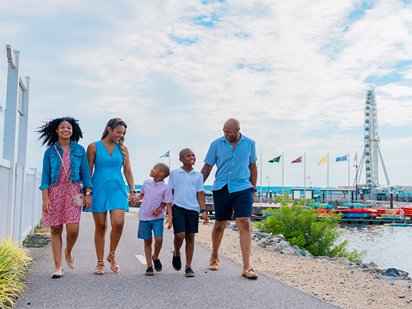 A family walks along a path by the water with a ferris wheel in the background. 