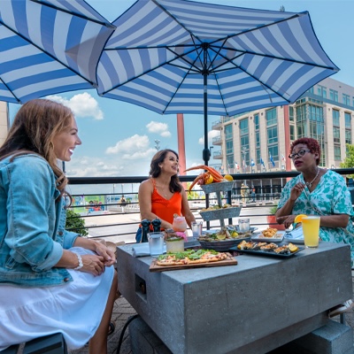 Three women chat and enjoy a seafood tower at a table on the water. 