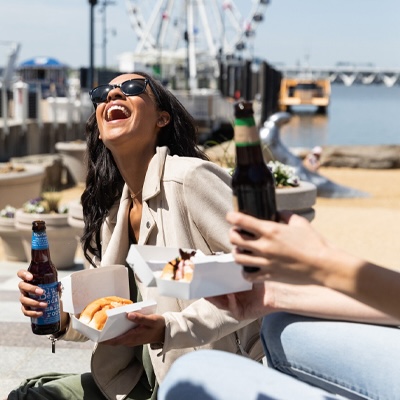 A woman in sunglasses laughs with a beer in her hand and the harbor in the background.
