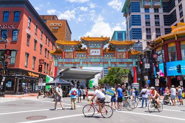 A lively street scene in Washington, DC's Chinatown shows people walking and biking beneath the colorful Friendship Archway during a community event.