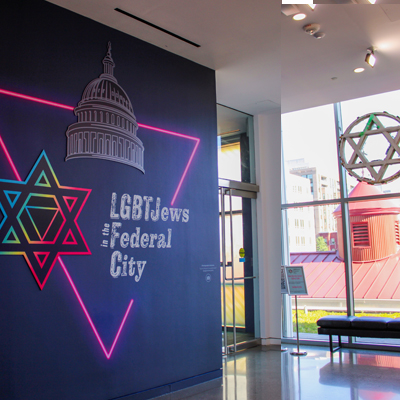 Photo of the LGBTJews in the Federal City exhibition entrance and title wall, featuring large illustration using the exhibition’s wordmark, an illustration of a neon pink triangle, a line drawing of the Capitol building and a colorful Jewish star with a pink triangle at its center.