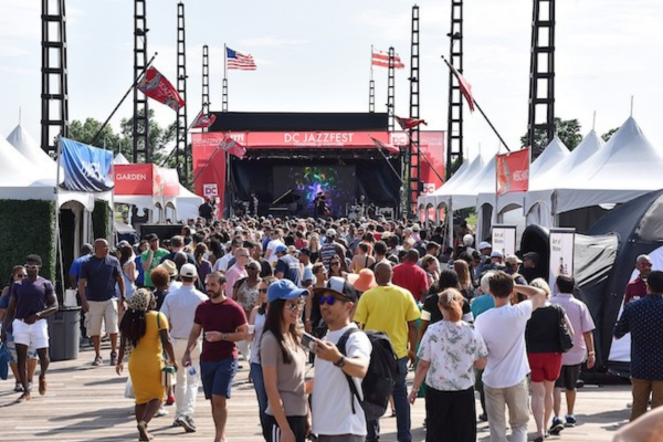 A large crowd gathers along the waterfront at DC JazzFest, heading toward a live performance stage under festival banners and flags.