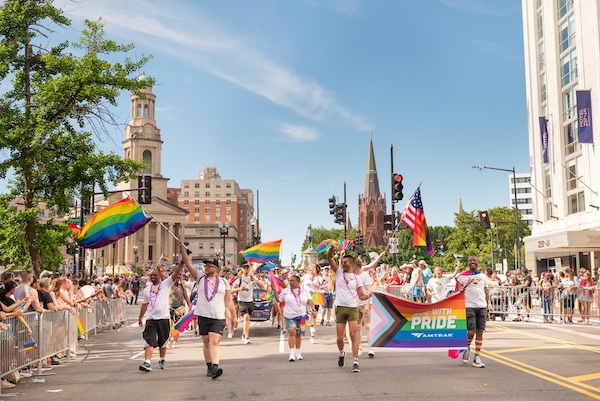 Parade participants wave Pride flags and carry a banner as they march down a sunny street lined with cheering spectators.