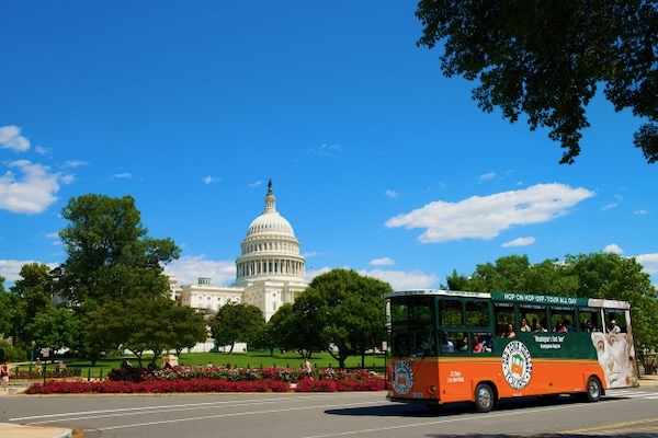 An orange and green Old Town Trolley bus drives past the U.S. Capitol building in Washington, D.C. on a sunny day. The Capitol dome rises behind trees and colorful flower beds under a bright blue sky with scattered clouds.