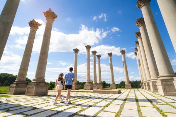 A couple walks among towering Corinthian columns under a bright blue sky at the National Arboretum in Washington, DC.