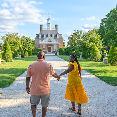 A couple holding hands walks along a gravel path toward a historic brick building with a white cupola, surrounded by manicured gardens and green trees under a bright blue sky with scattered clouds. The woman wears a yellow dress, and the man wears a peach shirt and gray shorts.