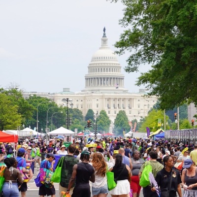 A vibrant crowd gathers for a street festival in Washington, D.C., with tents, food vendors, and people wearing colorful clothing and carrying tote bags. The U.S. Capitol building stands prominently in the background under a hazy sky.