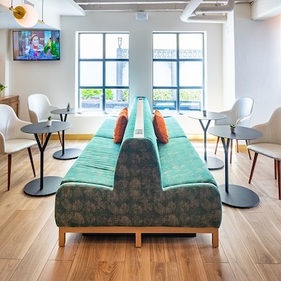 A bright, modern hotel sitting area with a central green bench, round tables, white chairs, and large windows letting in daylight.