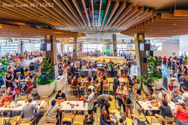 A vibrant crowd fills the spacious, modern food hall at La Cosecha, enjoying meals, drinks, and conversation.