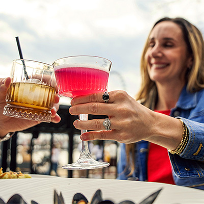 A woman smiling and holding a pink cocktail in her hand as she clinks her glass against another person’s glass.