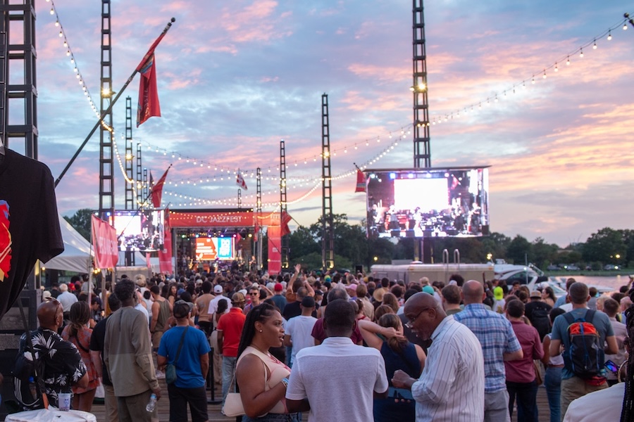 A large crowd gathers at DC JazzFest during sunset with string lights and video screens framing the stage.