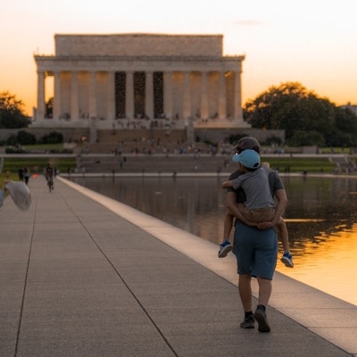 A man carries a child along the Reflecting Pool with the Lincoln Memorial in the background at dusk.
