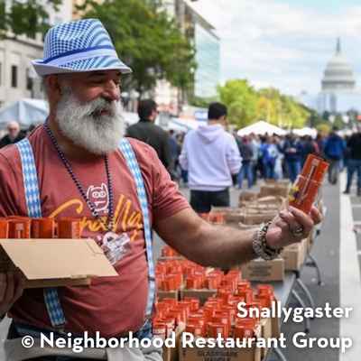 A person with a gray beard and blue hat handing out beer cans at Snallygaster festival with the U.S. Capitol in the background.