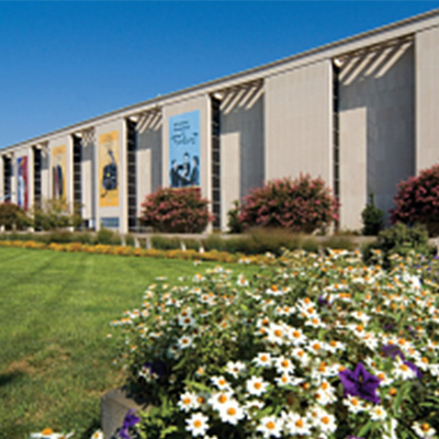 Front view of the National Museum of American History in Washington, DC with banners hanging on the facade, surrounded by blooming flowers and manicured lawns under a clear blue sky.