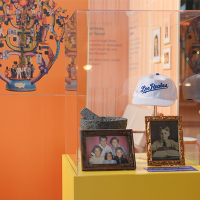 Exhibit case in the Making History pop-up exhibit featuring framed family photos, a white baseball cap embroidered with “Los Reales” and a volcanic stone mortar, set against a bright orange background with a colorful mixed-media artwork.