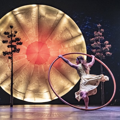 A Cirque du Soleil performer spins a large hoop on stage during Luzia in front of a glowing sun backdrop.