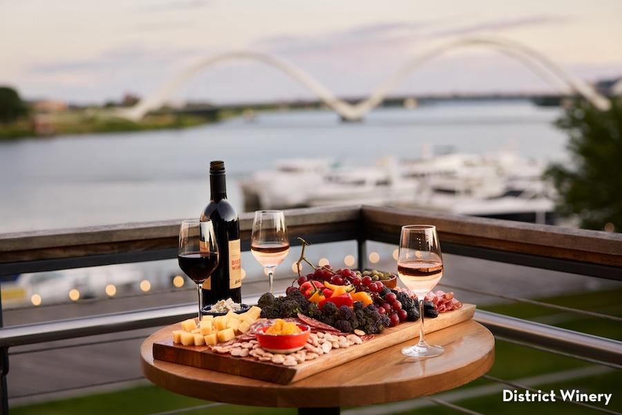 A charcuterie board and glasses of wine sit on a balcony overlooking the river and bridge at District Winery.